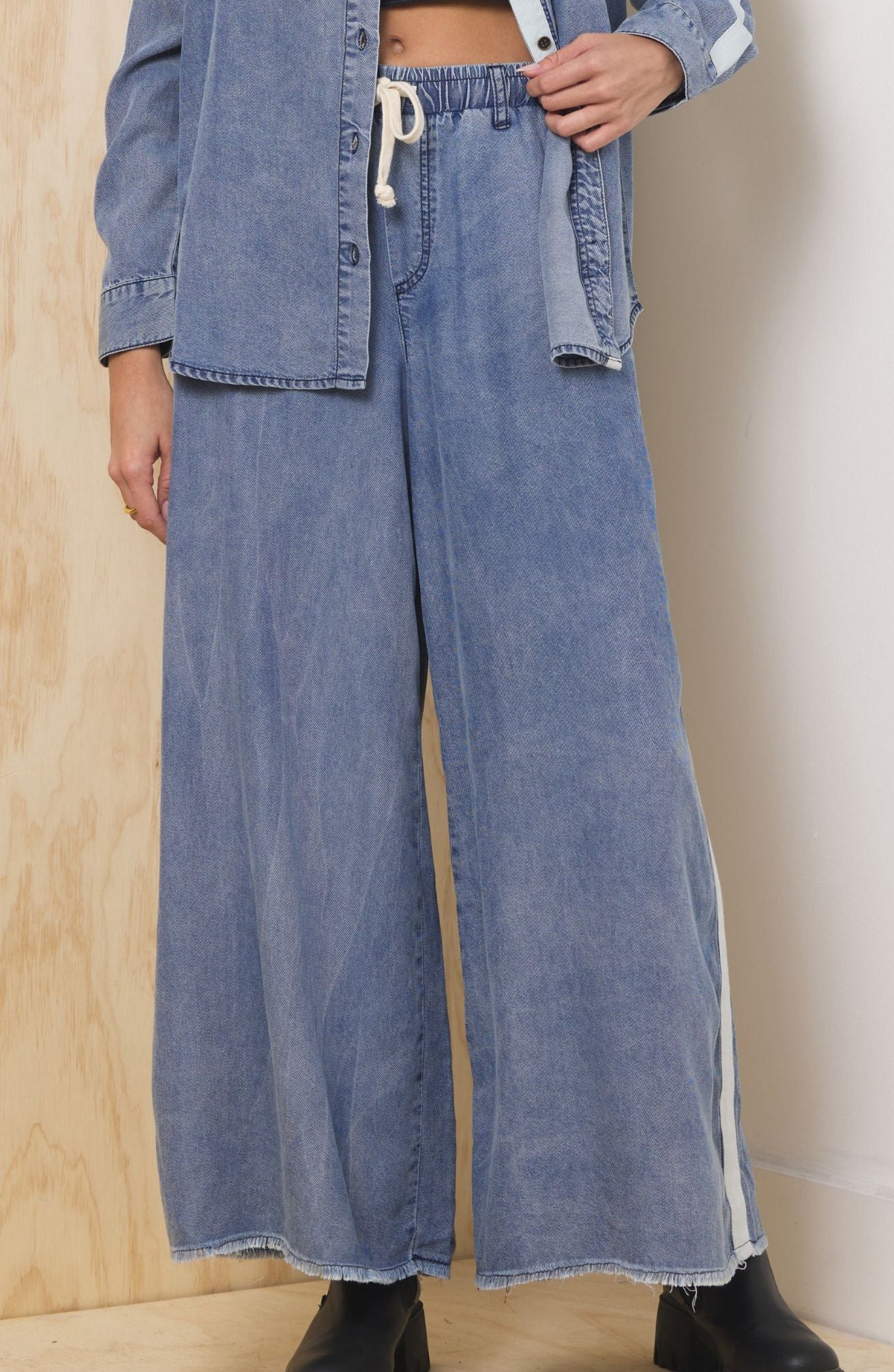 Woman wearing a denim outfit with wide-leg jeans and a matching jacket against a light-colored wall.