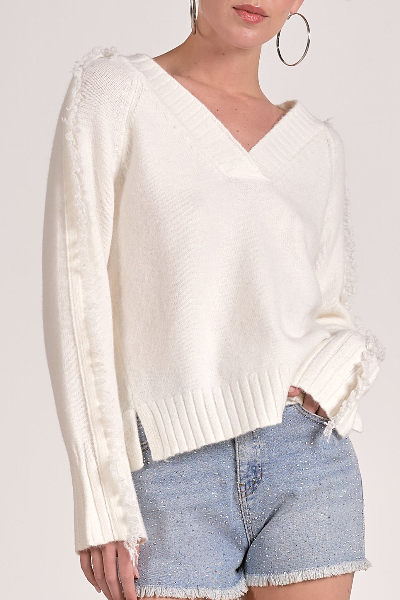 Model wearing a white v neck sweater with raw fringe detail down sleeves.